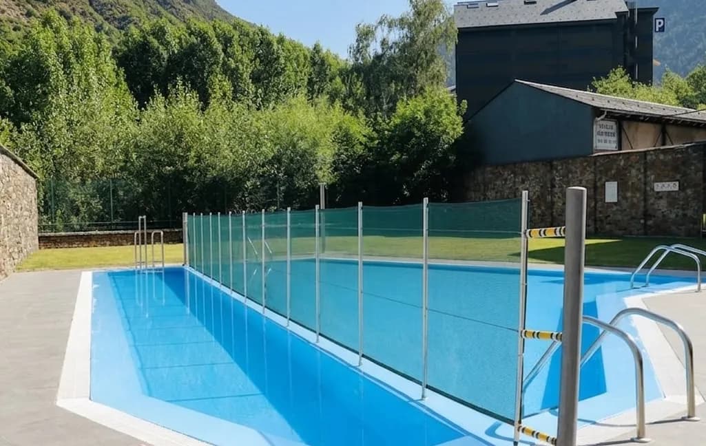 Una piscina exterior amb aigua blava clara, dividida per una tanca de malla verda. Al fons hi ha arbres verds frondosos i una muntanya, amb un edifici fosc a la dreta.