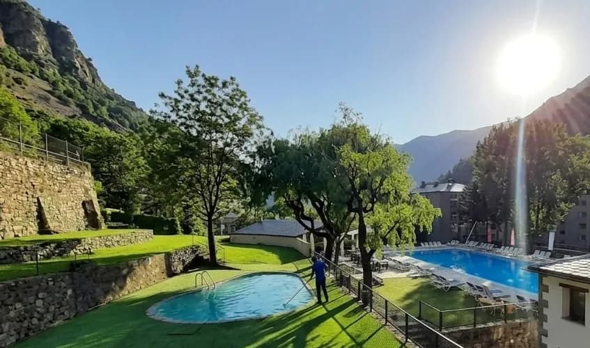 Una escena assolellada a l'aire lliure amb dues piscines, gespa verda i arbres, amb muntanyes de fons. Una persona està netejant la piscina més petita.