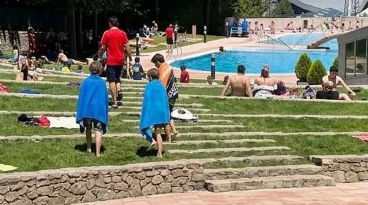 Dos nens embolicats amb tovalloles blaves pugen escales de gespa en una zona de piscina. Al fons, més persones es relaxen al voltant d'una piscina a l'aire lliure en un dia assolellat.