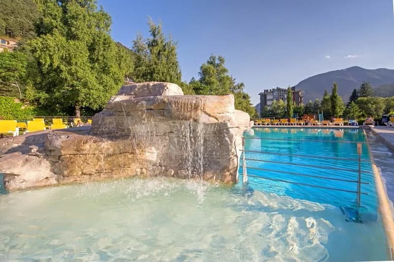 Una piscina exterior amb una cascada de roques en primer pla. Gandules grogues voregen la piscina, amb arbres verds, un edifici i muntanyes al fons sota un cel blau clar.