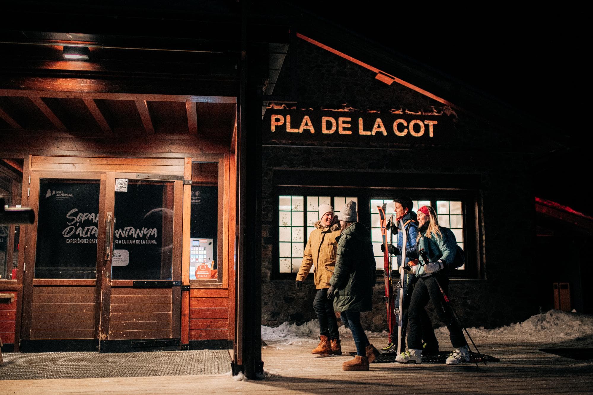 Quatre persones, algunes amb esquís i bastons, estan dempeus fora d'un edifici de fusta il·luminat càlidament amb neu a terra de nit. Un rètol prominent a l'edifici diu "PLA DE LA COT".