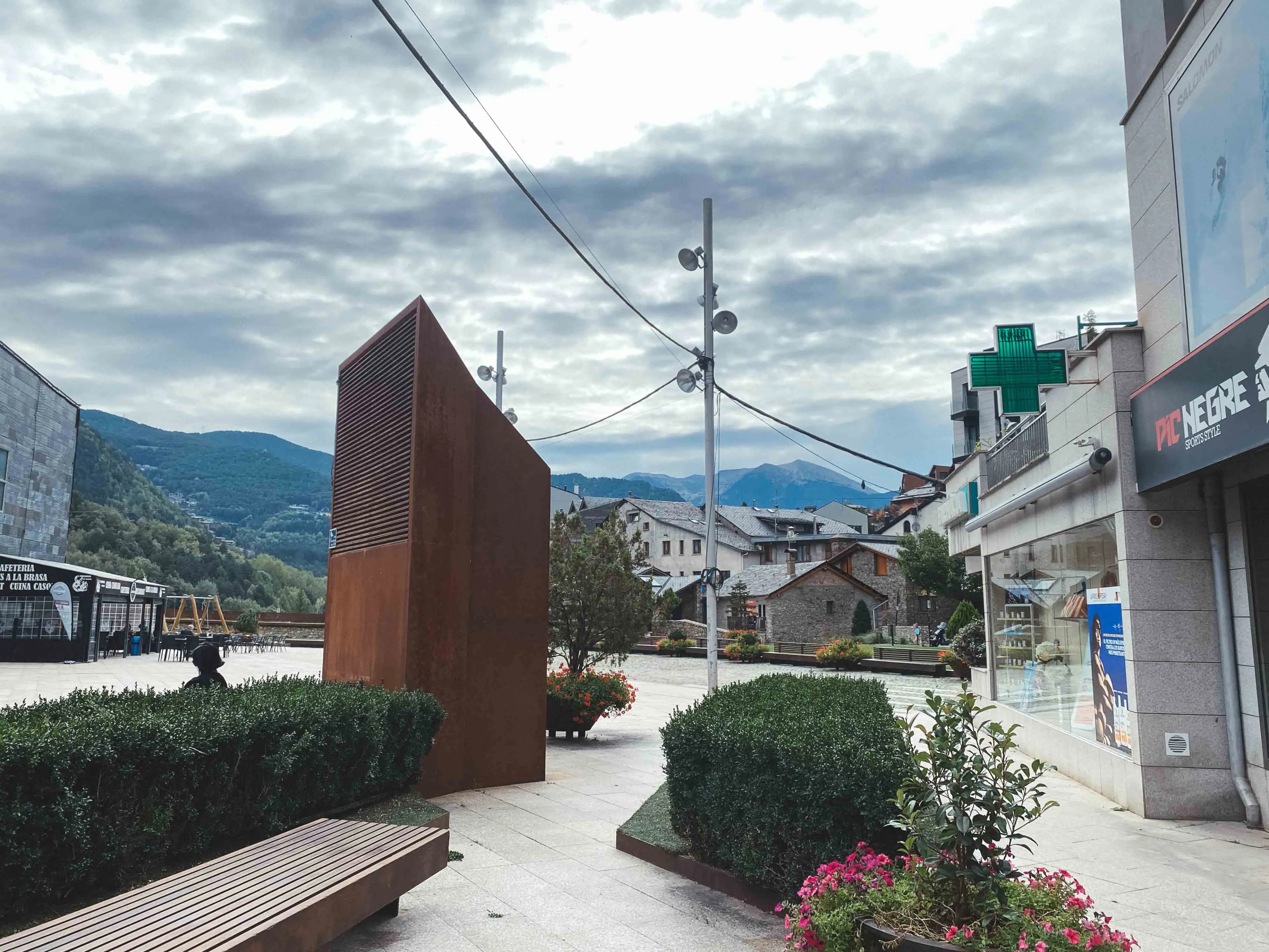 Una plaça de poble amb una estructura moderna de color rovell, arbustos verds i un banc de fusta en primer pla. Al fons, hi ha edificis tradicionals, una cafeteria, una farmàcia i muntanyes sota un cel ennuvolat.