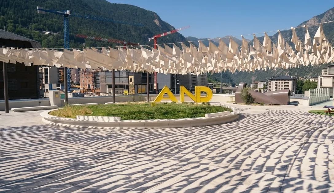 Una plaça pública amb una gran escultura groga 'AND' en un parterre circular d'herba. A dalt, banderes de tela blanca creen ombres estampades al terra pavimentat, amb edificis i muntanyes verdes al fons.