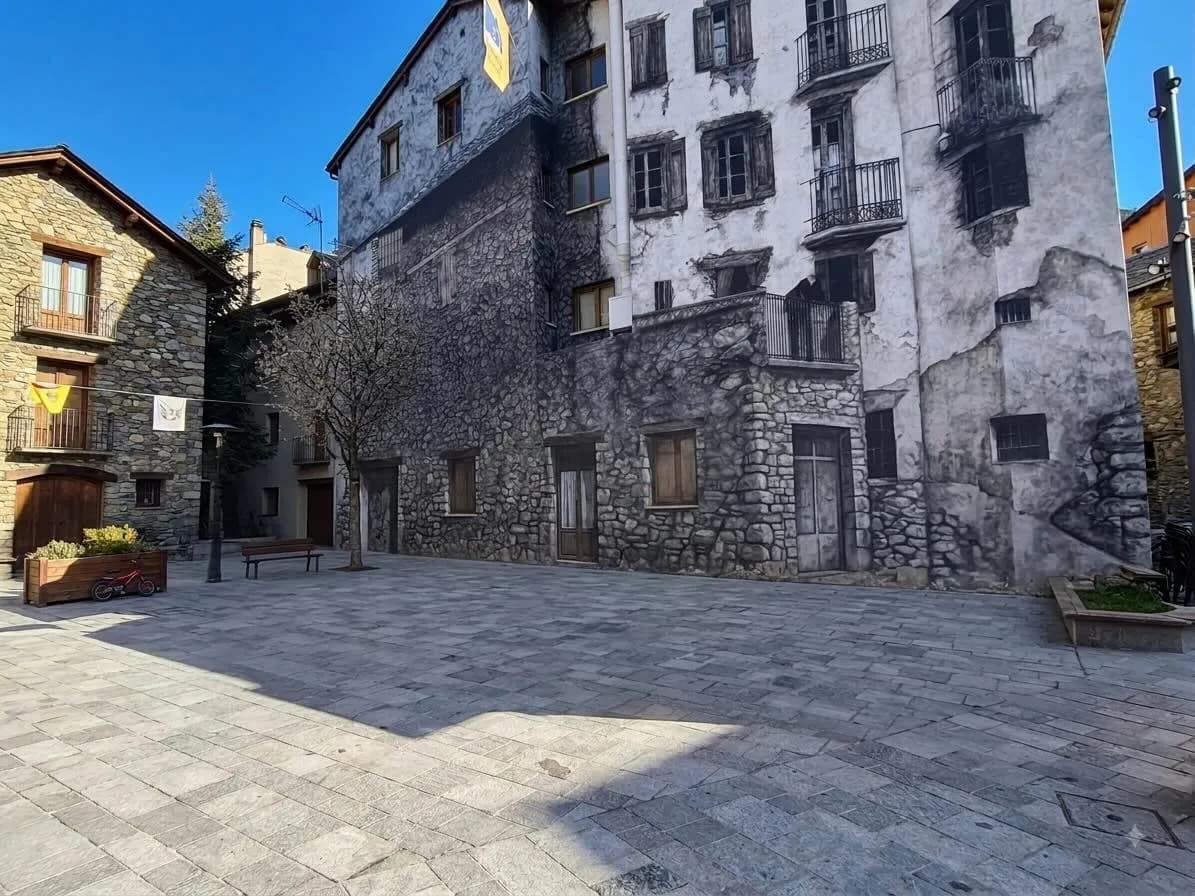 Una plaça de poble presenta un gran edifici amb un mural trampantojo d'una antiga façana de pedra, amb finestres i balcons, al costat d'un edifici de pedra real. La plaça està pavimentada amb llambordes grises i un arbre sense fulles es troba al centre.