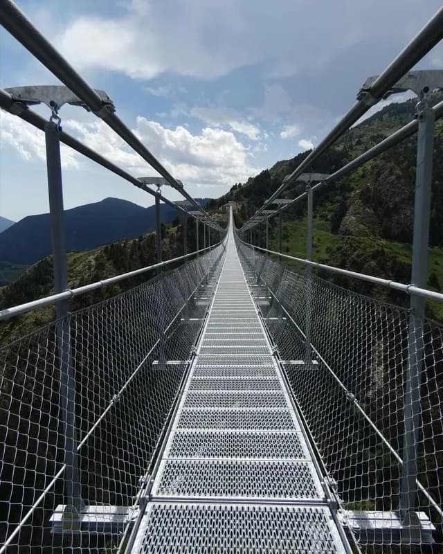 Un pont penjant llarg i estret amb una passarel·la de reixeta metàl·lica s'estén a la distància sobre una vall muntanyosa i verda sota un cel parcialment ennuvolat.
