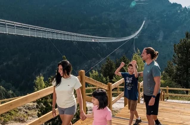 Una família de quatre, dos adults i dos nens, està dempeus en una plataforma de fusta, somrient i mirant cap a un pont penjant llarg i estret que s'estén per una vall muntanyosa boscosa al fons.