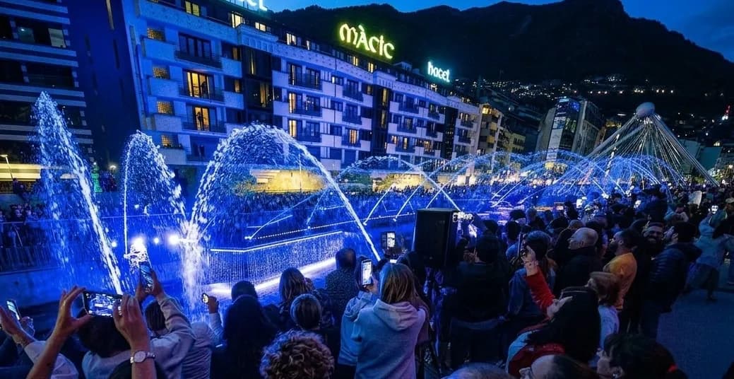 Una multitud de persones de nit observant i gravant fonts il·luminades de blau que ruixen aigua davant d'edificis, amb muntanyes al fons.