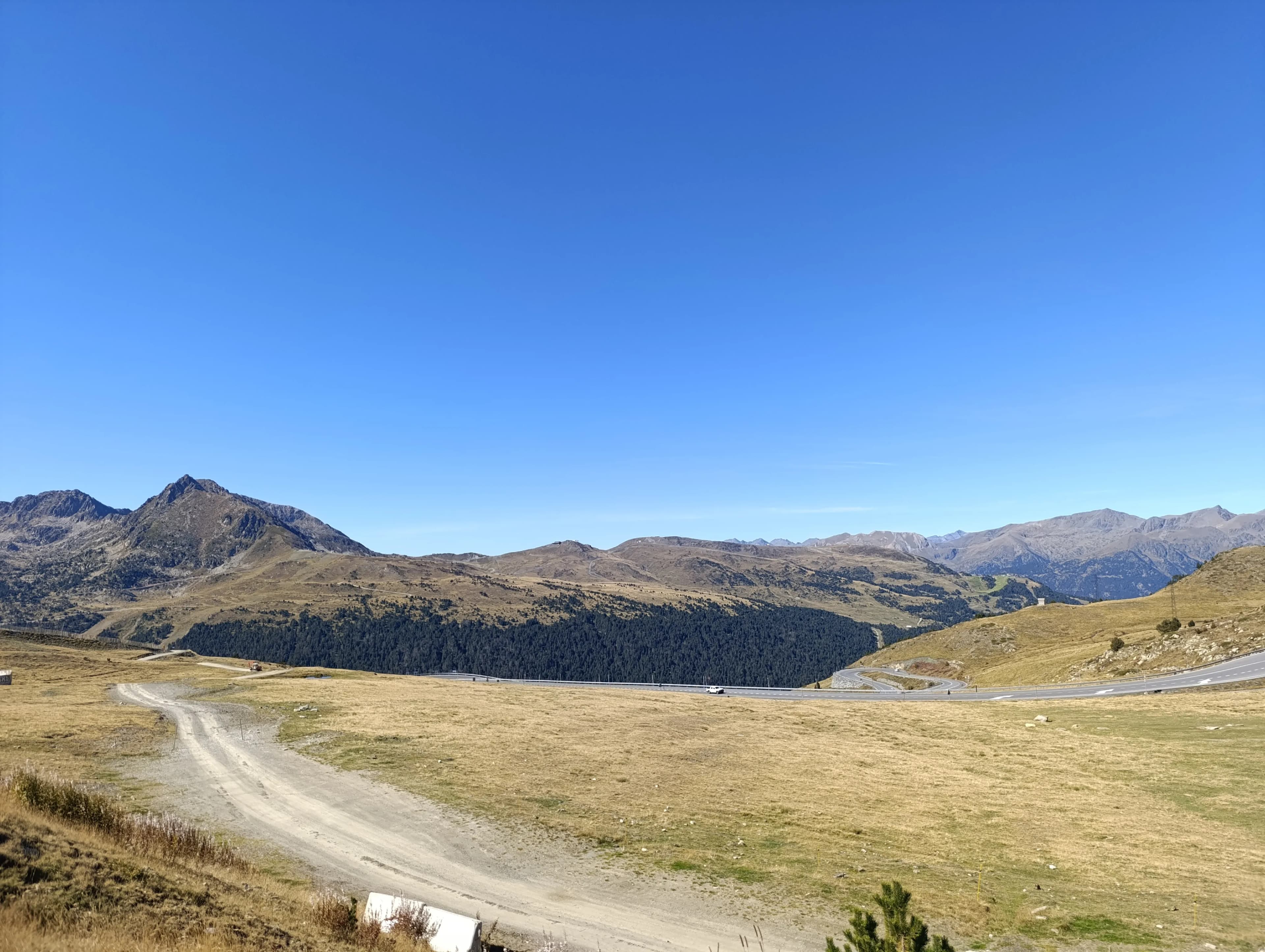 A wide panoramic view of a mountain landscape under a clear blue sky. A dirt road in the foreground leads to a winding paved road through a valley with golden grass and a dark forest, surrounded by rocky peaks.