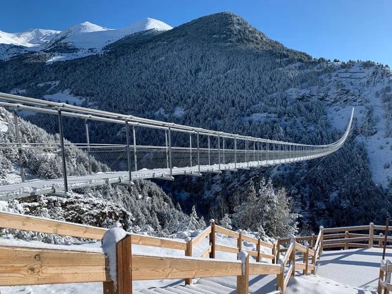 Un pont penjant llarg i estret s'estén sobre una vall coberta de neu. Muntanyes nevades i pins són visibles al fons sota un cel blau clar, amb una tanca de fusta en primer pla també coberta de neu.