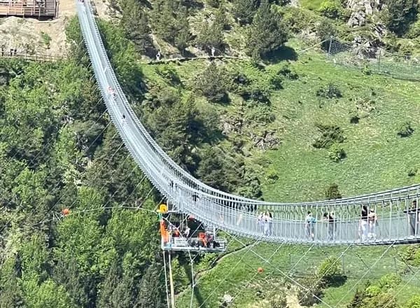 Un pont penjant llarg i estret, conegut com a pont tibetà, s'estén sobre una vall verda exuberant plena d'arbres. Hi ha persones caminant pel pont, i una plataforma a sota mostra una corda penjant, indicant una activitat de ràpel o puenting.