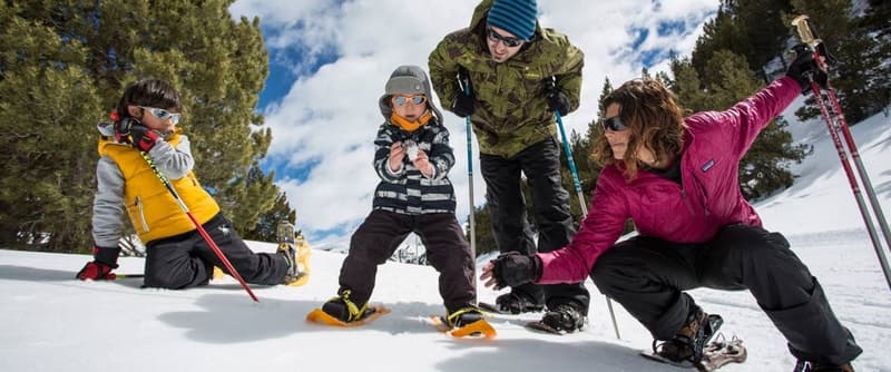 Una família de quatre, incloent dos adults i dos nens, fent raquetes de neu en un paisatge nevat i boscós. Un nen sosté una bola de neu mentre un altre s'agenolla a la neu.