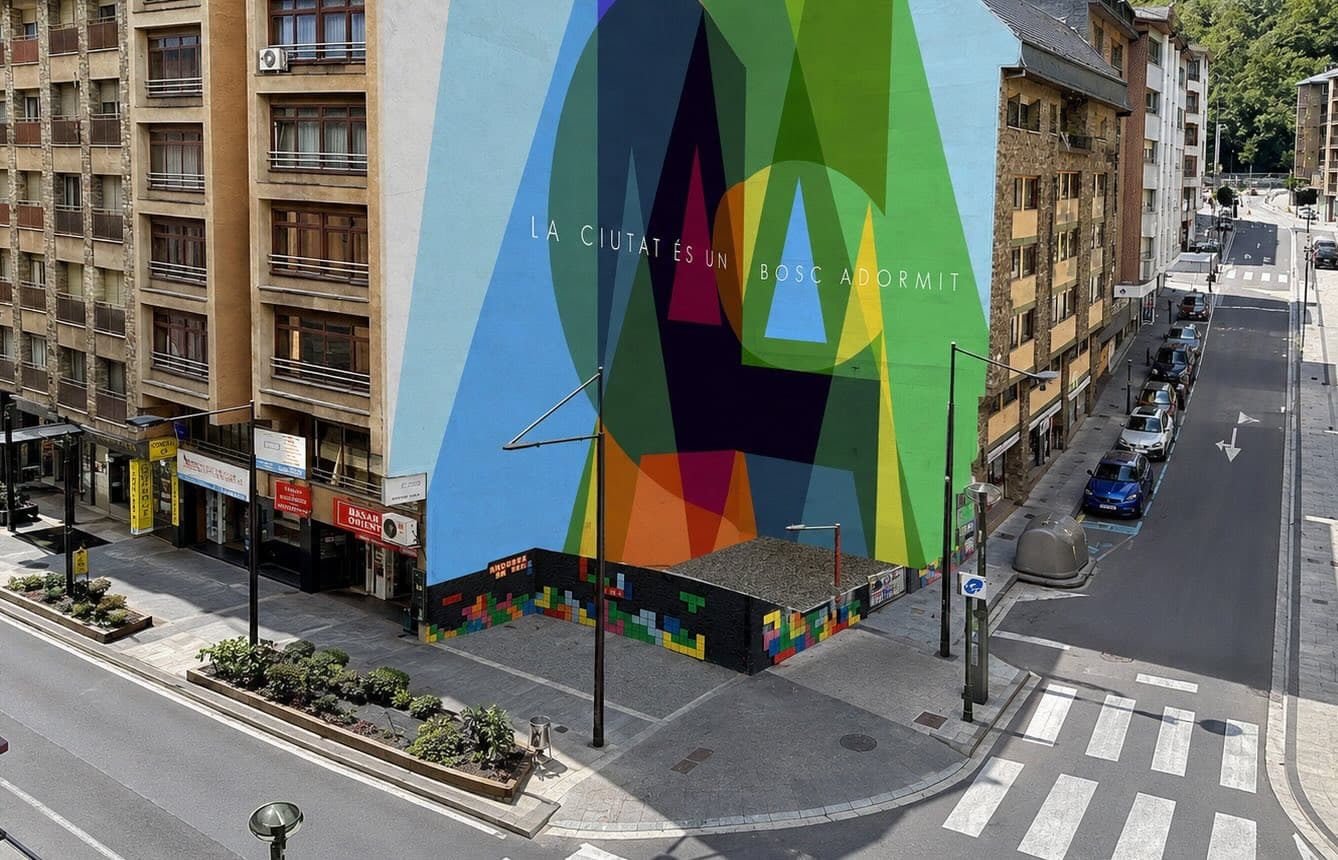 Vista aérea de una esquina de calle urbana con un gran edificio que presenta un vibrante mural geométrico que dice "LA CIUTAT ÉS UN BOSC ADORMIT". La calle de abajo tiene coches aparcados, pasos de peatones y un muro decorado con bloques coloridos tipo Tetris.