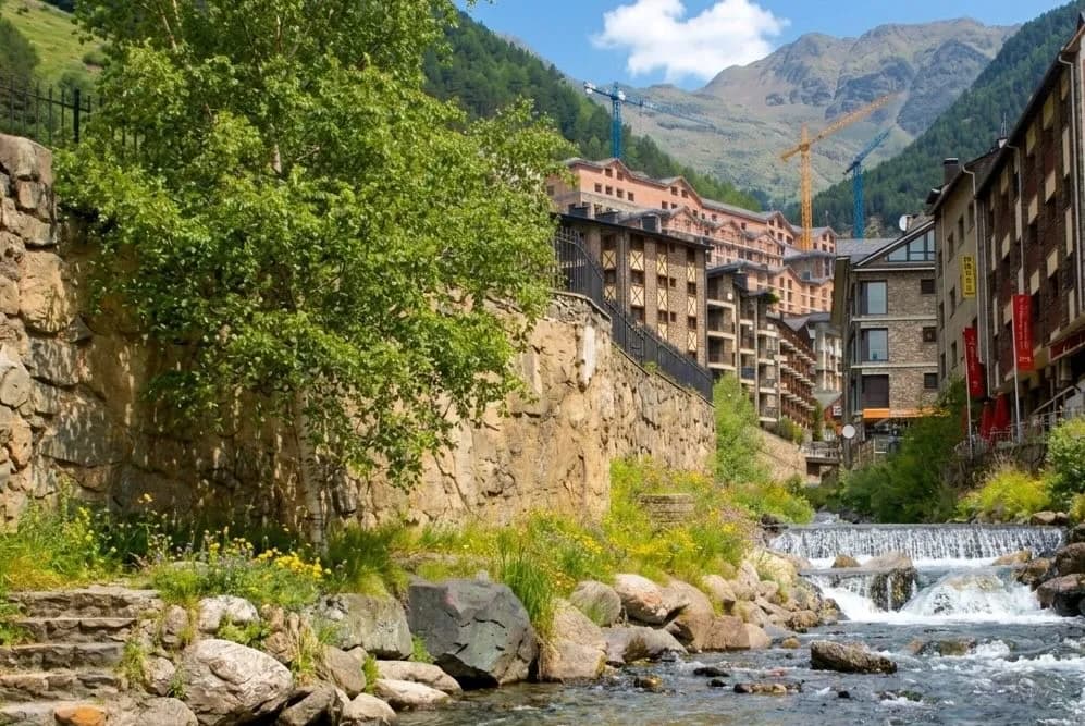 Un riu amb petites cascades flueix a través d'un poble de muntanya. Murs de pedra i arbres verds voregen la riba esquerra, mentre que edificis tradicionals s'alcen a la riba dreta, amb grues de construcció visibles contra les muntanyes boscoses al fons.