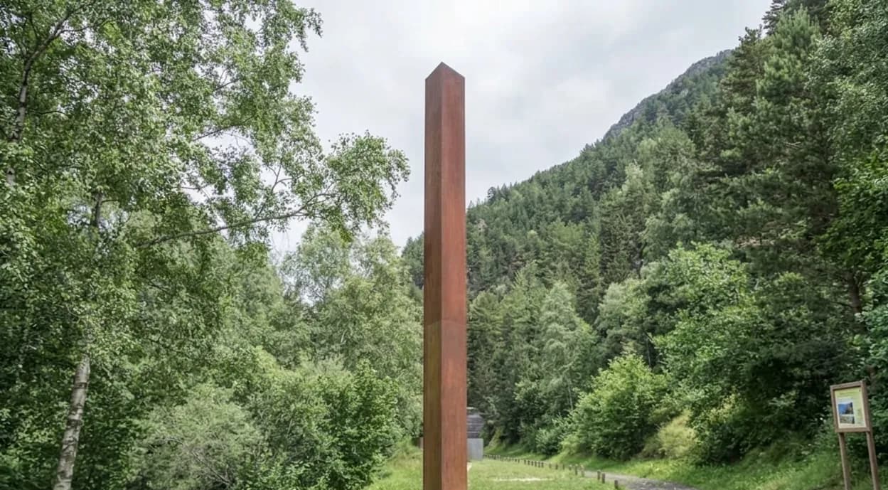 Un alt pilar rectangular de color marró rovellat s'alça enmig d'un dens bosc verd, amb una muntanya coberta d'arbres al fons sota un cel ennuvolat.