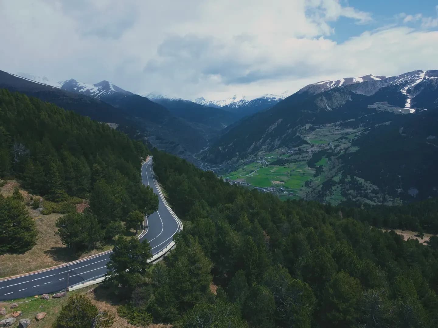 An aerial view of a winding road cutting through a dense pine forest on a mountain slope. In the distance, a green valley with a village is visible, framed by snow-capped mountains under a cloudy sky.