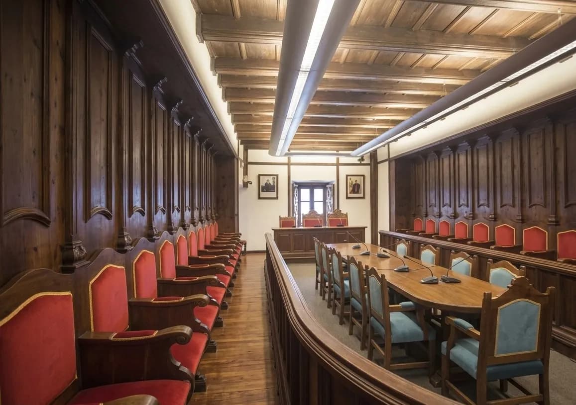Una sala formal amb panells de fusta fosca, una llarga taula de reunions amb cadires blaves i fileres de cadires vermelles al llarg de les parets. La sala té un sostre amb bigues i retrats a la paret del fons.