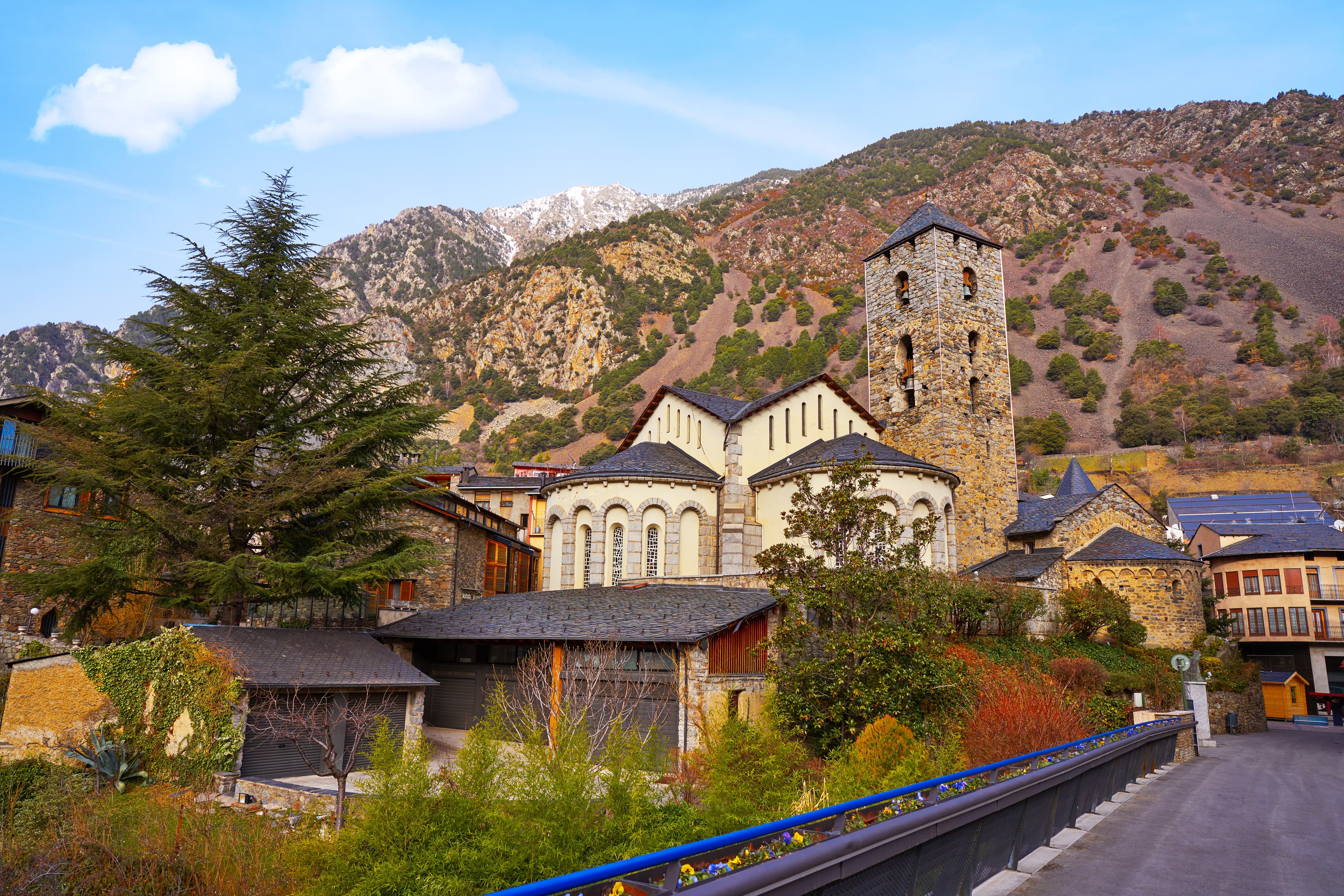 Una iglesia histórica de piedra con un alto campanario se alza entre edificios tradicionales en un pueblo de montaña. Picos nevados y laderas boscosas se elevan al fondo bajo un cielo azul, con un gran árbol de hoja perenne en primer plano.