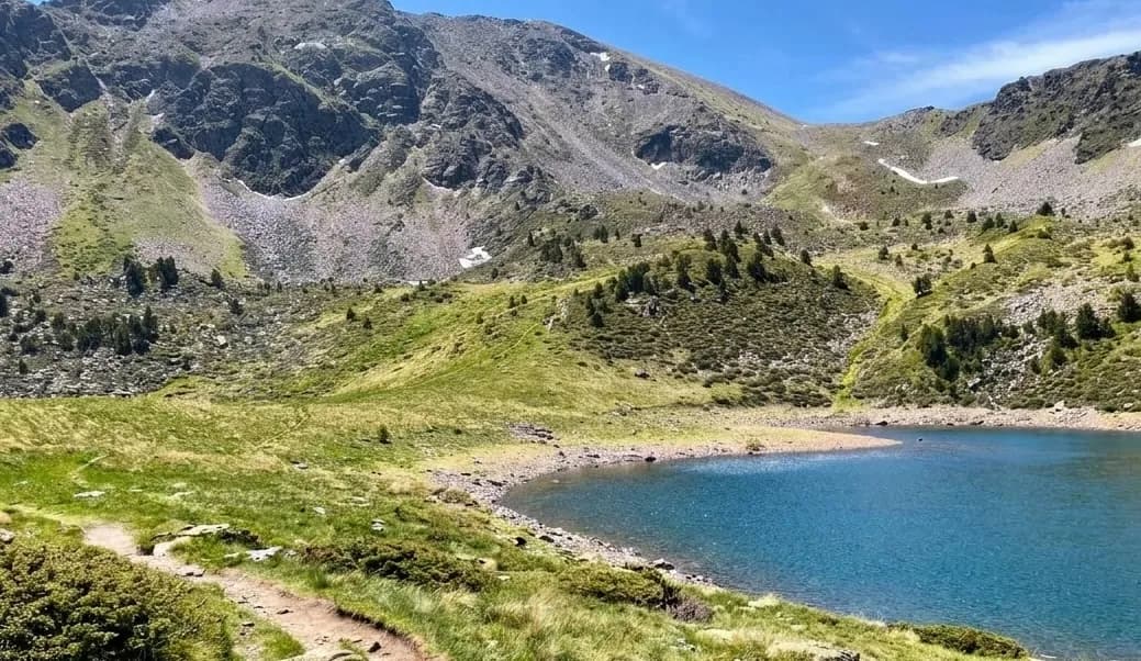 Un llac de muntanya blau clar envoltat de vessants verds i cims rocosos sota un cel brillant. Un camí de terra serpenteja pel costat esquerre del llac.