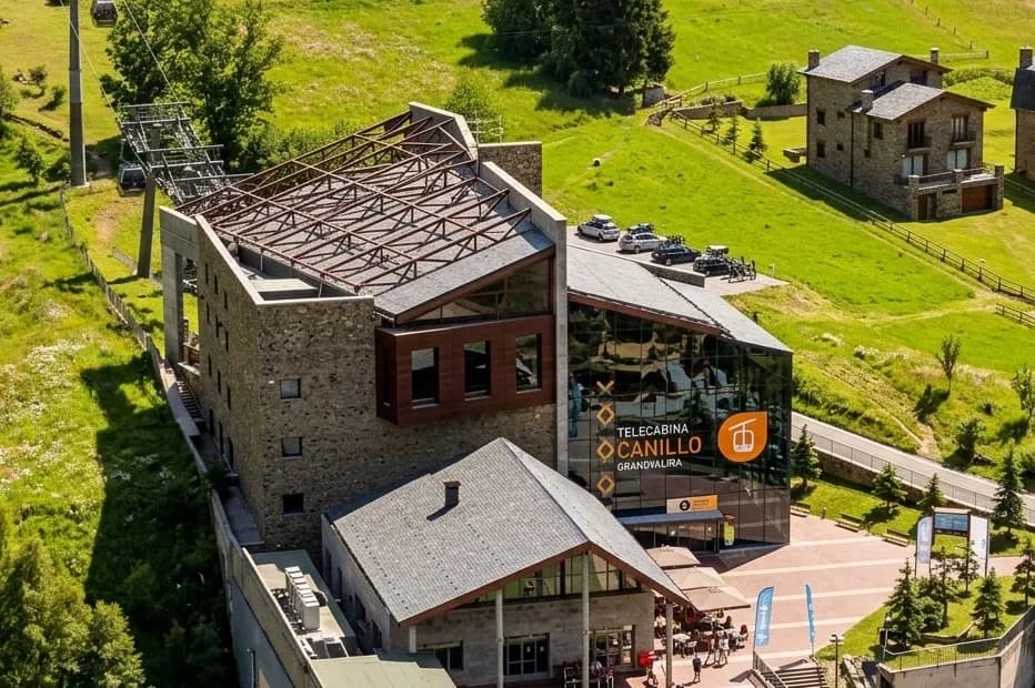 Vista aérea de la estación Telecabina Canillo Grandvalira, un edificio moderno con fachadas de piedra y cristal, ubicado en una ladera verde con cabinas de teleférico visibles en los cables.