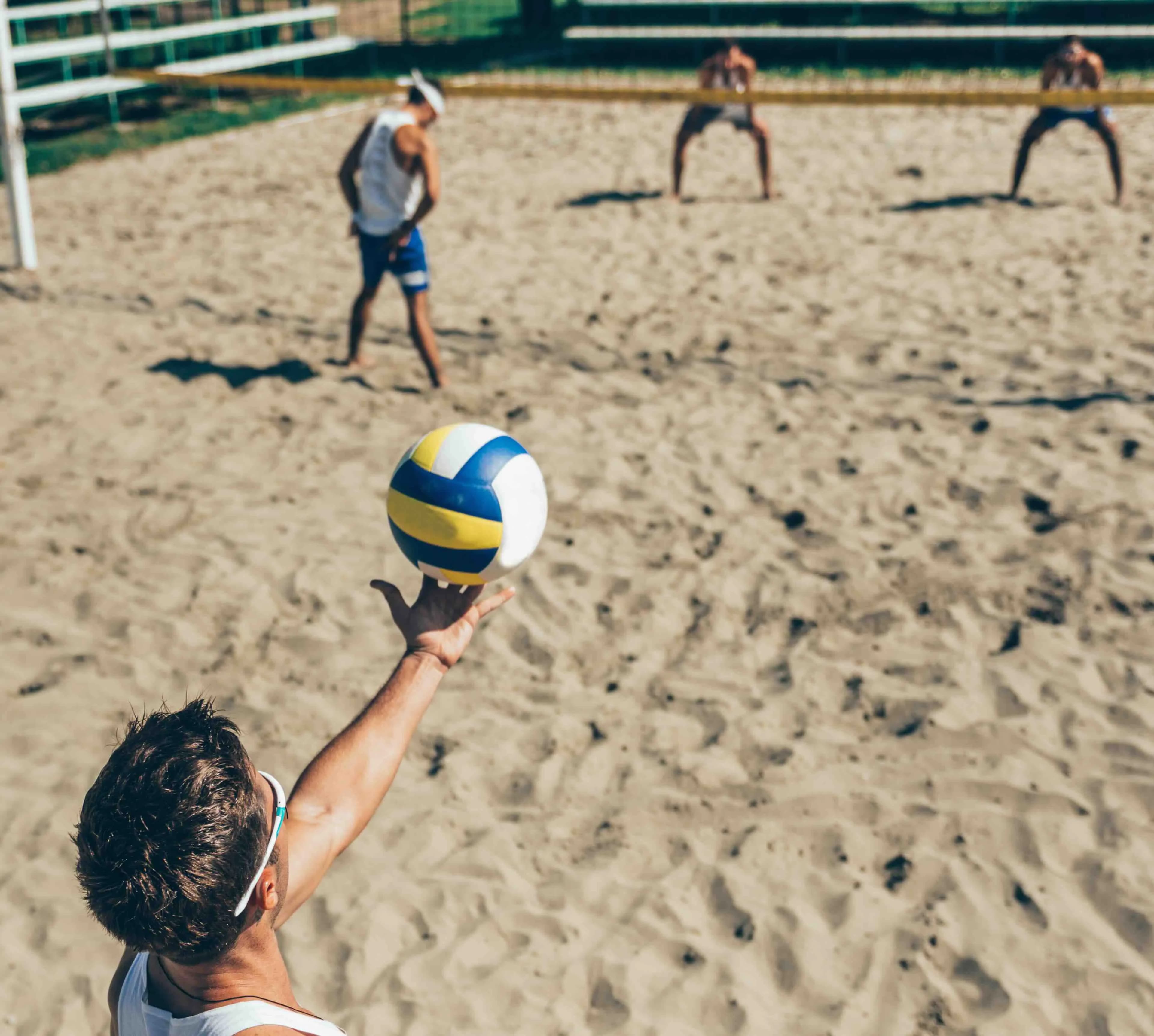 Un jugador de voleibol platja serveix la pilota, vist des de dalt, amb altres jugadors a la pista de sorra al fons.