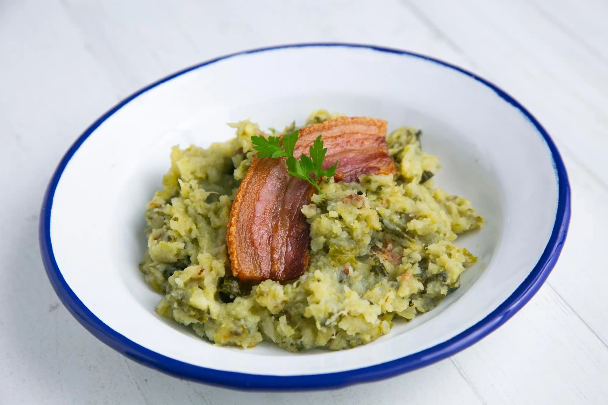 Un plat blanc amb vora blava conté una ració de Trinxat de Muntanya, un plat tradicional català de patates i col trinxades, coronat amb cansalada cruixent i una branqueta de julivert.