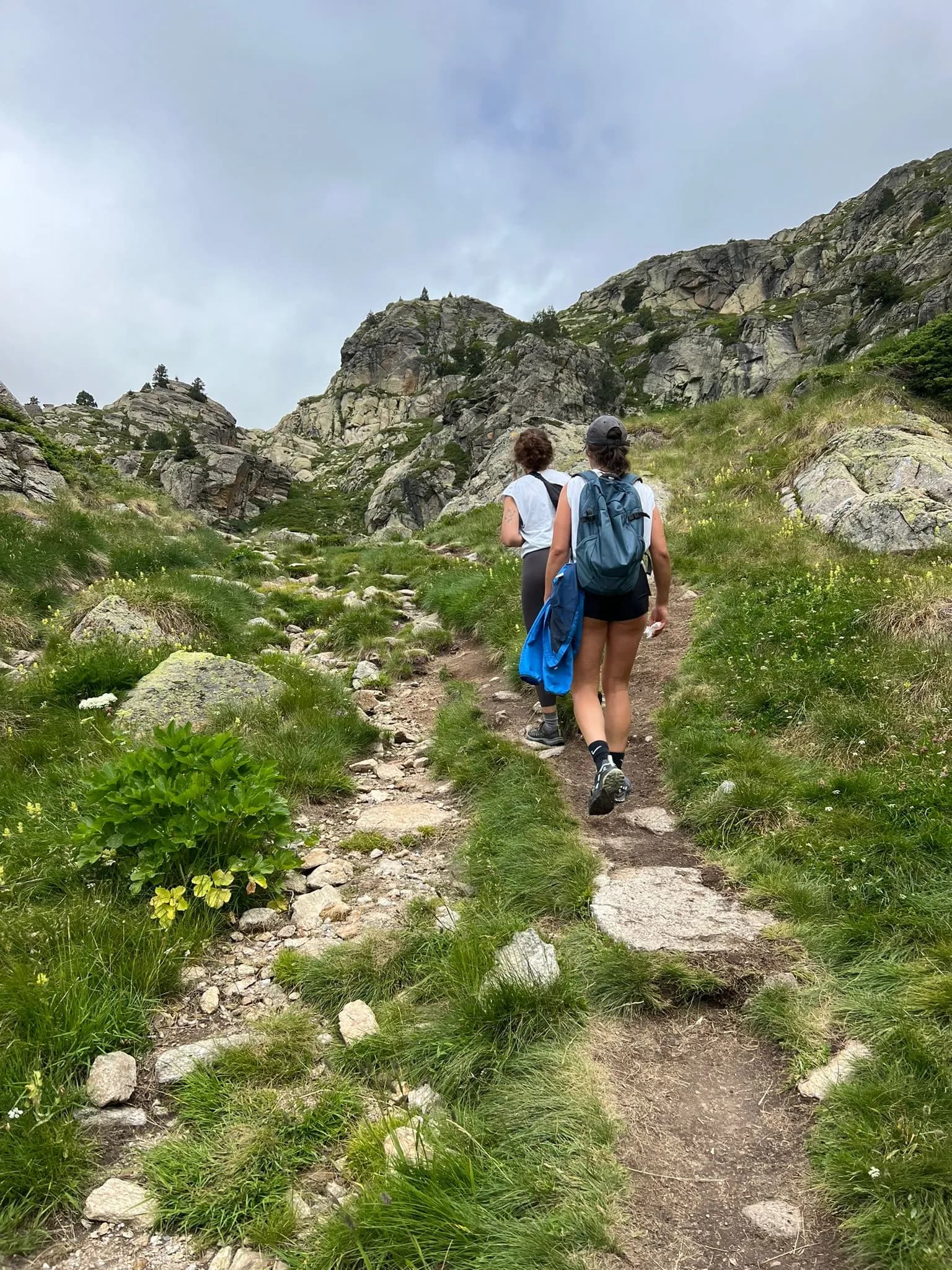 Dos excursionistes pugen per un camí de muntanya rocós i herbat. El camí està envoltat de vegetació verda i grans muntanyes escarpades sota un cel ennuvolat.