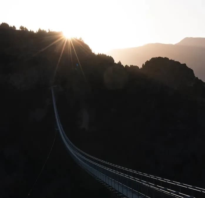 Un llarg pont penjant s'estén per una vall fosca, siluetejat contra una brillant explosió de sol sobre una muntanya boscosa a la sortida o posta de sol.