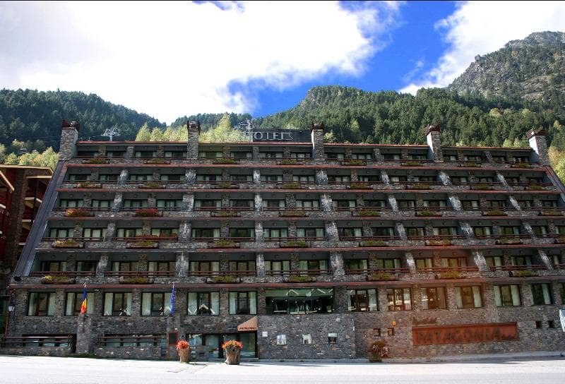 Un gran edificio de hotel de varias plantas con fachada de piedra y numerosos balcones con jardineras, enclavado en un paisaje de montañas boscosas bajo un cielo azul. El nombre 'PATAGONIA' es visible en la parte inferior derecha del edificio.