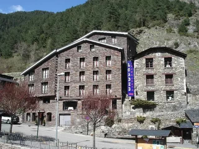 Edifici de diversos pisos construït en pedra fosca tradicional, amb finestres alineades i un cartell blau vertical que diu 'LOADED'. Es troba en un entorn de muntanya amb un vessant arbrat al fons i un carrer en primer pla.
