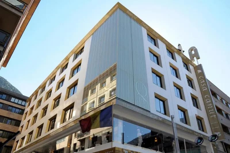 Un modern edifici d'hotel de diverses plantes amb façanes blanques i daurades, amb nombroses finestres i un rètol prominent que diu "a casa canut". La cantonada té vidre reflectant, mostrant el reflex d'un edifici adjacent.