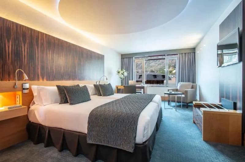 Una moderna habitació d'hotel amb un llit gran, un escriptori al costat de la finestra i vistes a les muntanyes. L'habitació presenta una decoració contemporània amb una paret de capçal de fusta fosca i catifa blava clara.