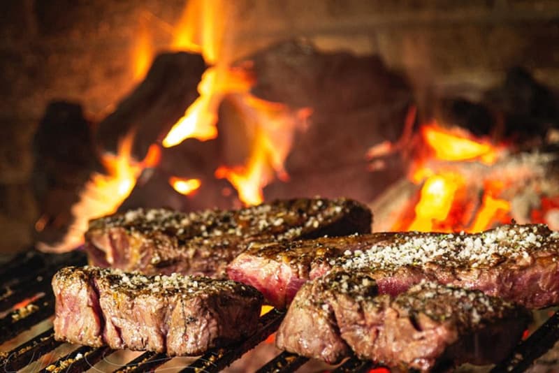 Plusieurs steaks assaisonnés grillent sur un feu ouvert, avec des flammes vives visibles en arrière-plan.