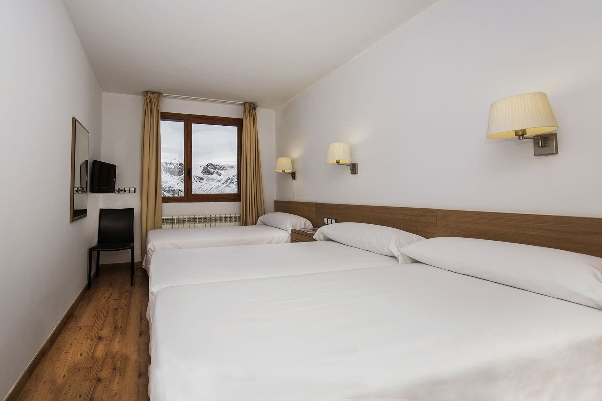 Una habitación de hotel con tres camas individuales vestidas con ropa de cama blanca y suelo de madera. Una ventana con marcos de madera y cortinas beige ofrece vistas a montañas nevadas.