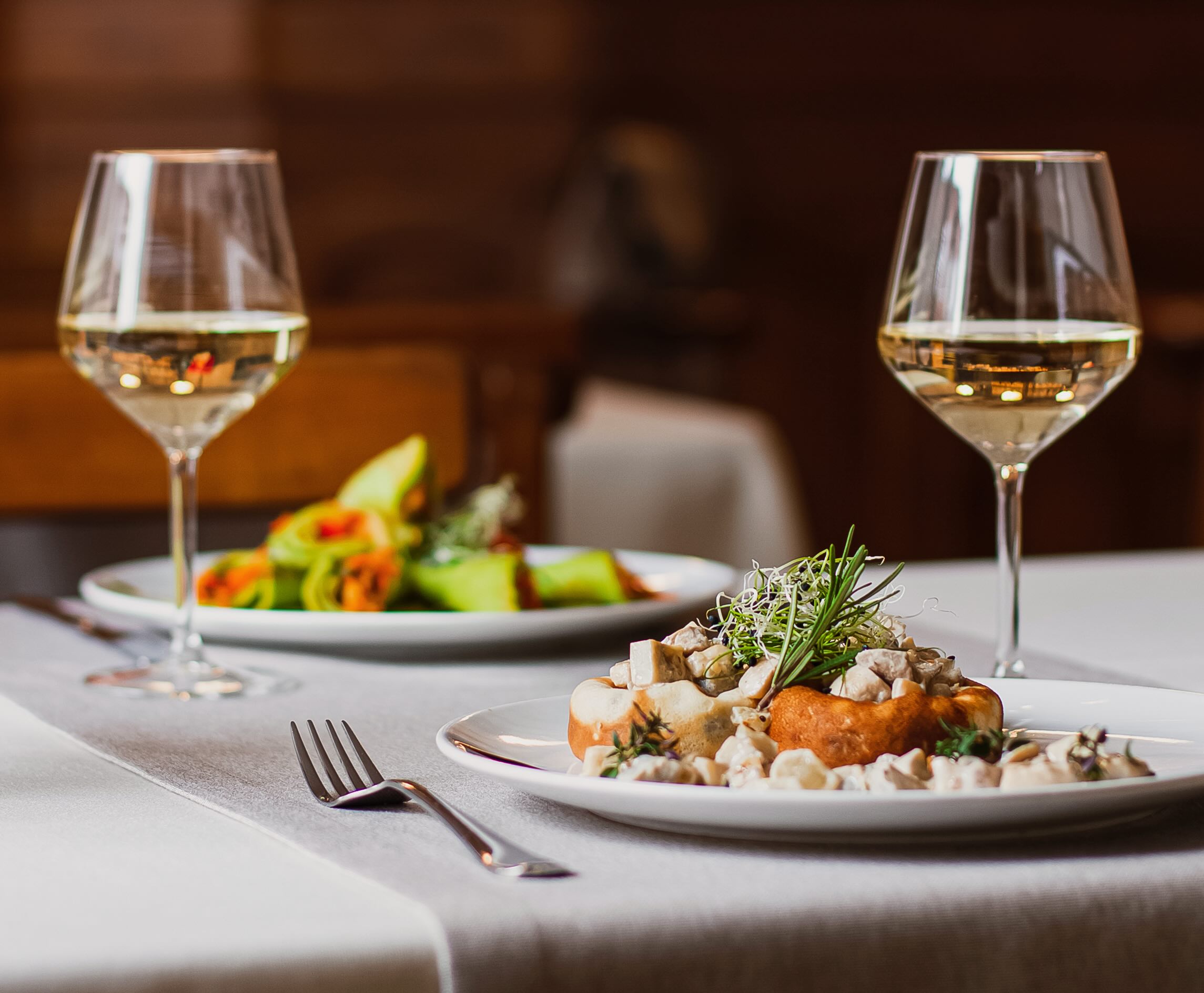 Dos elegantes platos de comida y dos copas de vino blanco están dispuestos sobre un mantel blanco. El plato en primer plano presenta champiñones, un pastel y hierbas frescas, mientras que el del fondo muestra rollos verdes.