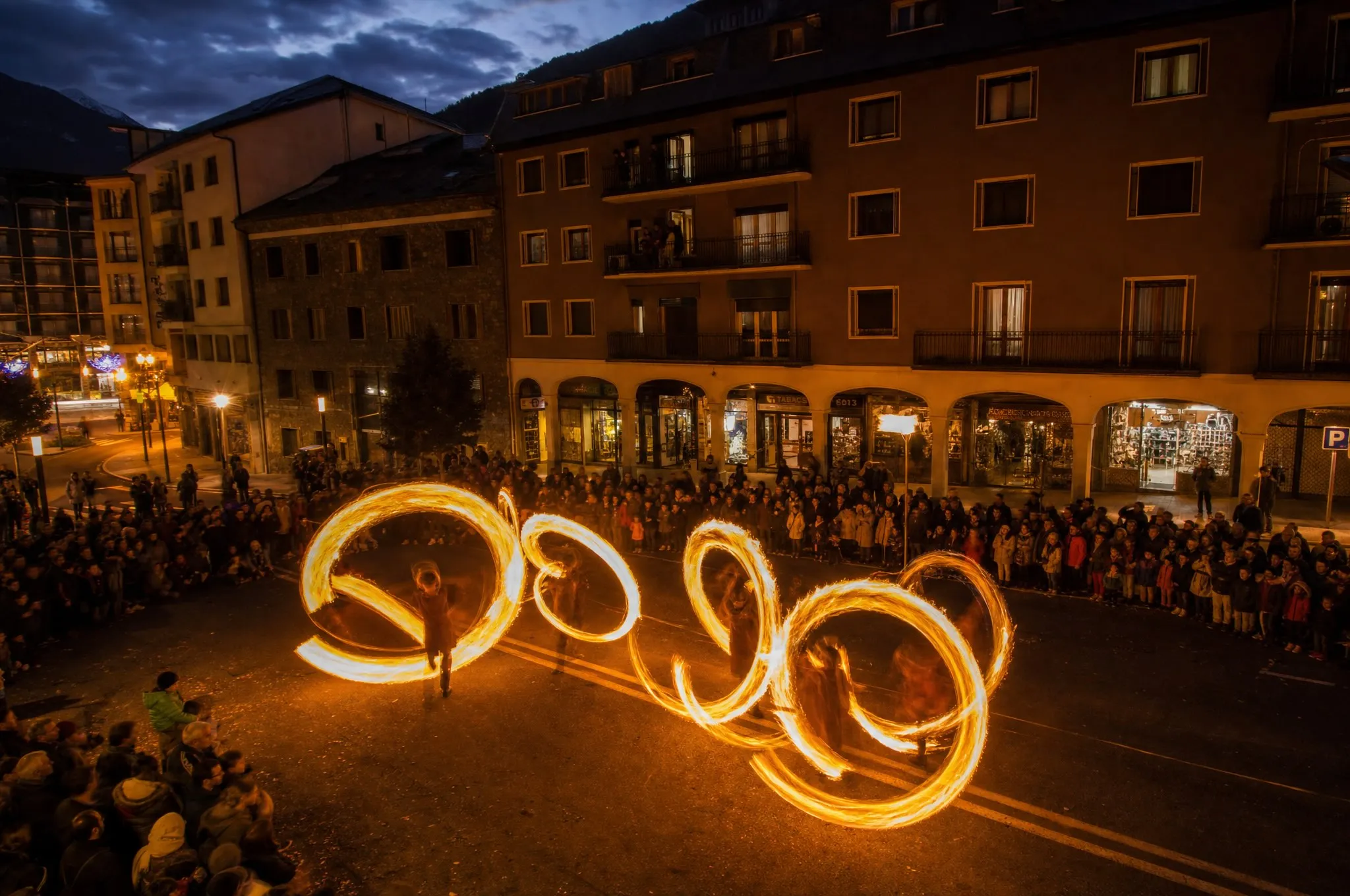 Artistas crean brillantes rastros de fuego giratorios en una calle por la noche, observados por una multitud de personas a lo largo de la calle. Edificios con ventanas iluminadas son visibles al fondo bajo un cielo oscuro.
