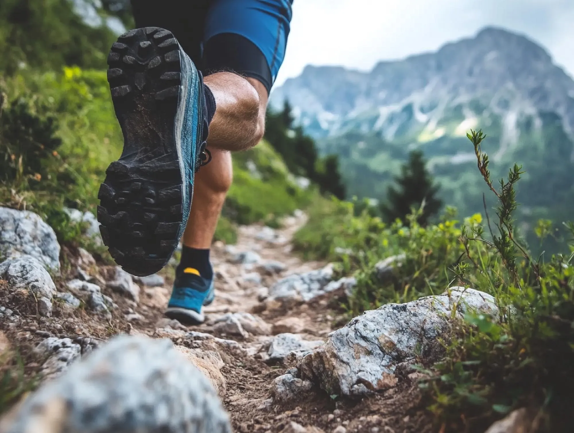 Gros plan de la jambe et du pied d'une personne en chaussures de course bleues, en pleine foulée sur un sentier de montagne rocheux. Des collines vertes et des montagnes sont visibles en arrière-plan.