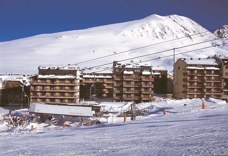 Una pista de esquí con una estación de telesilla en primer plano, que se dirige hacia varios edificios cubiertos de nieve y una gran montaña blanca bajo un cielo azul claro.