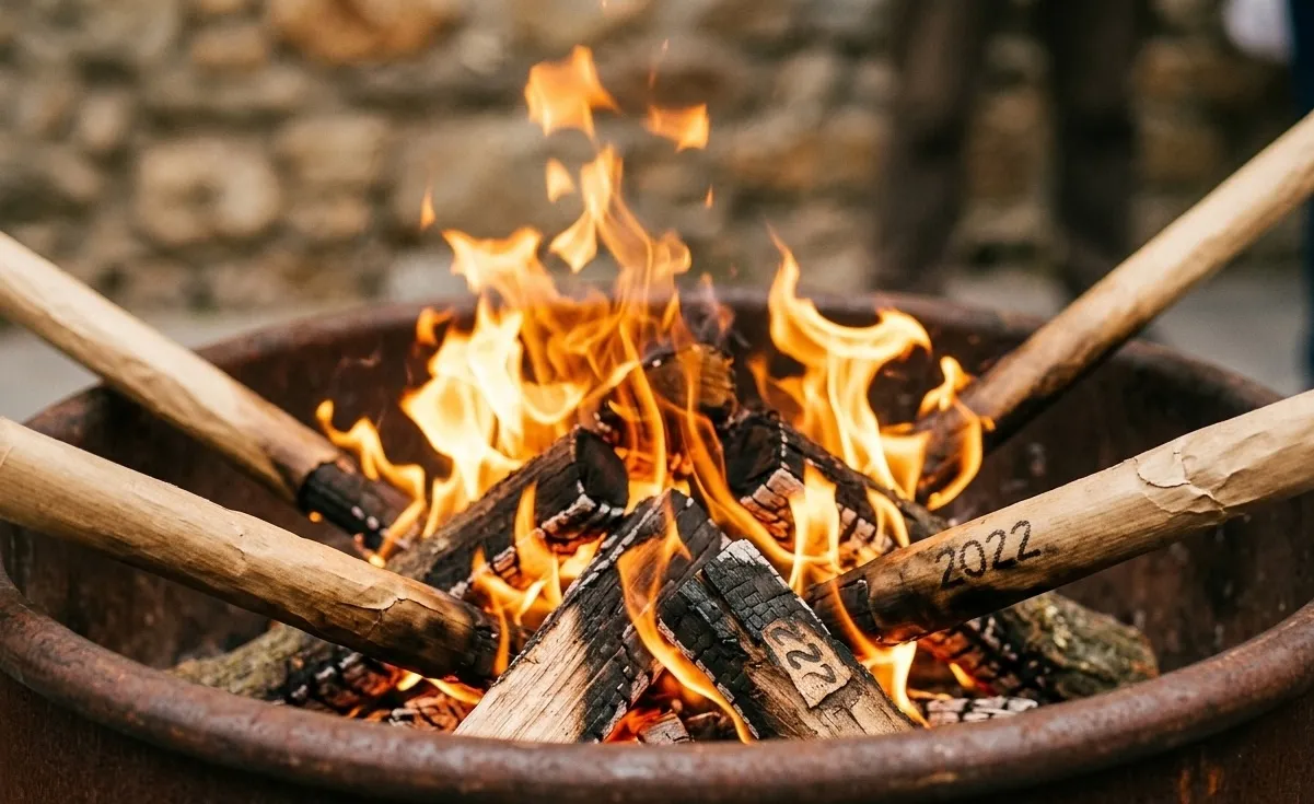 Un primer plano de una hoguera en un brasero de metal, con llamas naranjas brillantes consumiendo leños. Varios palos de madera, algunos marcados con '2022', están colocados alrededor del fuego.